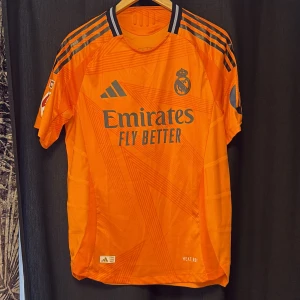 Real Madrid orange målvaktströja Adidas - Säljer en officiell Real Madrid målvaktströja från Adidas i orange med svarta detaljer och tryck. Tröjan har korta ärmar, klubbmärke och sponsortryck på bröstet samt autenticitetsmärke. Tillverkad i lätt och ventilerande funktionsmaterial. Tröjan är äkta och är köpt i Madrid.
