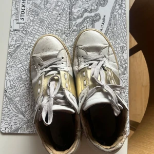 Valentino open - Snygga sneakers i vitt och guld med klassisk snörning och rund tå. Skorna har en platt sula och detaljer i både vitt och metallic guld, vilket ger dem en unik look. Perfekta för dig som vill sticka ut lite extra i vardagen. Ganska slitna där av priset. 