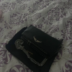 Svart axelväska Zadig & Voltaire - Säljer en svart axelväska från Zadig & Voltaire i skinn med coola silverdetaljer, kedjerem och dubbla dragkedjor. Väskan har nitar längs kanterna och en ikonisk vinglogga framtill. Perfekt för dig som gillar edgy och stilren design.
