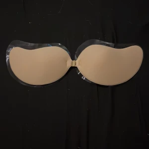 Beige bandeau tubtopp från SHEIN - Säljer en beige bandeau tubtopp från SHEIN i storlek A. Toppen är axelbandslös och har en slät, formad design med transparenta kanter och knäppning framtill. Perfekt under plagg med öppen rygg eller axelbandslöst.