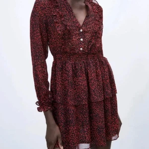 Röd och svart mönstrad kortklänning - Supersöt kortklänning i rött och svart med små leopardmönster, volangdetaljer och knappar framtill. Klänningen har långärmade ärmar med rynk vid ärmslut och v-ringning. Perfekt för dig som gillar trendiga och lekfulla plagg.