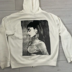 Vit Ariana Grande hoodie med tryck - Vit hoodie med dragkedja och huva, framtill står det 'Ariana Grande' i svart text. På ryggen finns ett stort svartvitt tryck av en kvinna i profil. Hoodien har känguruficka och är i mjukt material, perfekt för dig som gillar stilrena merch-plagg. Skicka för prisförslag mm