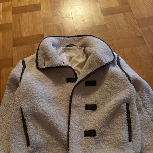 teddyjacka hm  - Säljer en fluffig vit teddyjacka med svarta kantband och snygga knäppningar framtill. Jackan har stor krage och två fickor med svarta detaljer. Perfekt för dig som vill ha en mysig och trendig look under kyliga dagar.