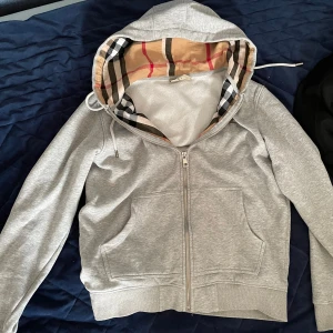 Grå Burberry hoodie med klassiskt rutmönster - Snygg grå hoodie från Burberry med dragkedja och huva. Insidan av huvan har det ikoniska Burberry-rutmönstret i beige, svart, vitt och rött. storlek m men passar som s