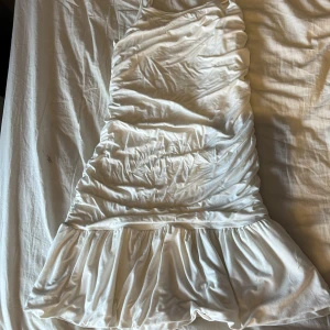 Vit mini-klänning med volang - Supersöt vit kortklänning med smala axelband och volang nertill. Klänningen har en figurnära passform och är lätt rynkad för en snygg effekt. Perfekt för sommarens alla tillfällen!