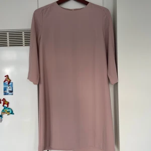 Ljusrosa klänning från Femme Collection - Enkel och stilren ljusrosa klänning från Femme Collection. Modellen är rak med trekvartsärm och rund halsringning. Klänningen har dragkedja bak och är tillverkad i ett lätt, slätt tyg som ger en clean look.