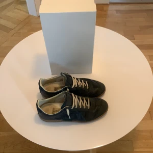 Blå Maison Margiela  - Maison Margiela sneakers i svart skinn och mocka med beige snörning och innersida. Klassisk rund tå och platt gummisula i naturfärg. Snygg minimalistisk design, levereras med originalkartong och dustbags.