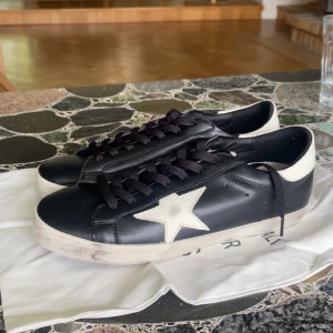 Golden Goose svarta sneakers Storlek 42 - Golden Goose sneakers i svart skinn med vit stjärndetalj på sidan och vit häl med logotyp. Skorna har svarta snören och en vit, något smutsad sula för en cool, använd look. Klassisk rund tå och låg modell. Perfekta för dig som gillar streetstyle. Kommer utan Skokartong