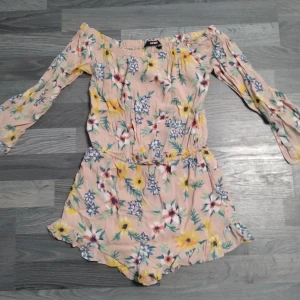 Blommig shortsdress med volang från Bik Bok - Supersöt shortsdress från Bik Bok i ljusrosa med blommigt mönster i gult, vitt, grönt och lila. Modellen har volangkant nertill och resår i midjan för en bekväm passform. Perfekta för sommarens alla äventyr!