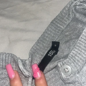 Grå kofta från 157 med knappar - En basic grå kofta från 157 i storlek M. Koftan är ribbstickad, har långa ärmar och stängs med flera knappar framtill. Perfekt att slänga över en t-shirt eller linne när det är lite kyligare ute.