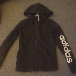 Svart Adidas hoodie med tryck - Svart hoodie från Adidas med vit logga längs ena ärmen. Tröjan har huva, dragkedja framtill och två fickor. Perfekt för en sportig look eller chill dagar. Klassisk och enkel design som funkar till det mesta.
