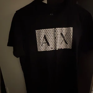 Svart t-shirt från Armani Exchange - Svart t-shirt från Armani Exchange med stort grafiskt A|X-tryck i vitt framtill. Klassisk passform med rund hals och korta ärmar. Materialet känns mjukt och skönt, perfekt till jeans eller shorts. Diskret logga i nacken.
