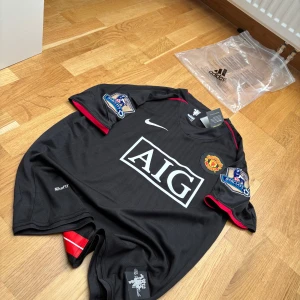 Manchester United Ronaldo 7 Nike tröja - Svart Manchester United fotbollströja med Ronaldo 7 på ryggen. Tröjan har röda detaljer, Premier League-patchar på ärmarna och AIG som sponsor på bröstet. Tillverkad i NikeFIT Dry-material som andas, perfekt för match eller träning.