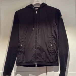 Moncler Cardigan svart - Svart cardigan från Moncler med dragkedja framtill och två fickor med knappar. Den är storlek L men passar M bra.  Jackan är i nyskick inte använd mycket. Säljes pga den enbart samlar damm i garderoben. Den har ribbade muddar, huva med dragsko och Moncler-logga på ärmen. Snygga ränder på huvan ger en sportig touch. Perfekt för dig som gillar stilren design. Hör av dig vid frågor!