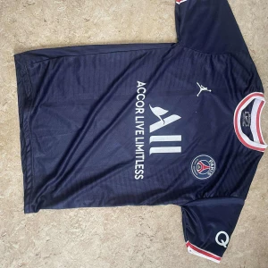 PSG Messi 30 matchtröja Jordan - Snygg mörkblå PSG fotbollströja med Messi 30 på ryggen. Tröjan har vita och röda detaljer vid krage och ärmslut, Paris Saint-Germain-logga på bröstet och Jordan-logga. Tillverkad i lätt och ventilerande material, perfekt för fotbollsplanen eller att bära som supporter.