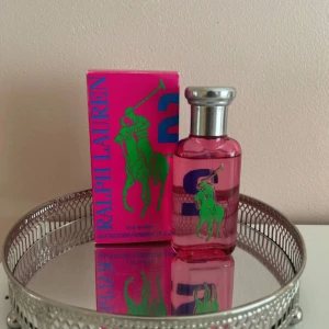 Ralph Lauren Big Pony 2 parfym - Fräsch och färgstark parfym från Ralph Lauren, Big Pony 2 för kvinnor. Flaskan är rosa med silverfärgad kork och har en grön polospelare på framsidan. Kommer i en snygg rosa kartong med blå och gröna detaljer. Perfekt för dig som gillar lekfulla och energiska dofter.