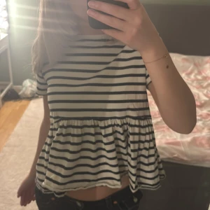 Randig volangblus i bomull - Supersöt blus med marinblå och vita ränder, rund hals och korta ärmar. Modellen är lite croppad med volang nedtill som ger en avslappnad och trendig look. Perfekt för dig som gillar en chill men ändå stylish vibe.