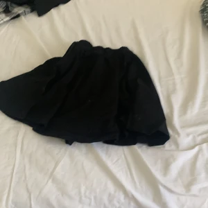 Svart kortkjol med volang - En svart kortkjol med volang och elastisk midja. Kjolen har en flowy och luftig känsla, perfekt för att skapa en cool och avslappnad look. Passar till sneakers eller boots för en trendig stil.