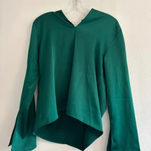 Grön omlottopp från H&M - Snygg grön omlottopp från H&M med lång ärm och coola slitsar vid ärmsluten. Toppen har en asymmetrisk nederkant och v-ringad hals. Perfekt för dig som gillar unika detaljer och vill sticka ut med en modern look.