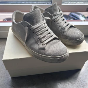 Gråa sneakers från Axel Arigato i mocka - Snygga grå sneakers från Axel Arigato i mjuk mocka med klassisk låg profil och rund tå. Skorna har platt sula, diskret logga på sidan och matchande grå snörning. Perfekta för en clean och stilren look.