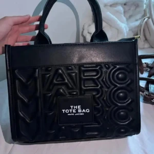 Svart tote bag - Svart handväska från Marc Jacobs med präglad logotyp över hela väskan och tydlig patch framtill med texten 'The Tote Bag'. Väskan har två stadiga handtag och är tillverkad i syntetskinn. 💕