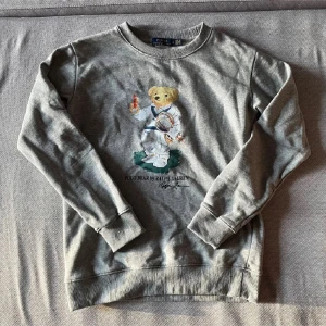 Grå sweatshirt Polo Bear Ralph Lauren - Grå sweatshirt från Polo Ralph Lauren med ikoniska Polo Bear-trycket på bröstet. Tröjan har rund hals, långa ärmar och ribbade muddar. Tillverkad i mjuk bomullsmix, perfekt för en chill och sportig vibe.