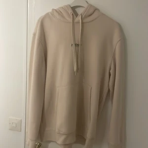 Beige hoodie från Artisan - Snygg beige hoodie från Artisan med minimalistisk svart text på bröstet. Tröjan har huva med snörning, stor känguruficka och långa ärmar. Perfekt för en chill och avslappnad stil. Mjuk och skön, troligtvis i bomull eller polyesterblandning.
