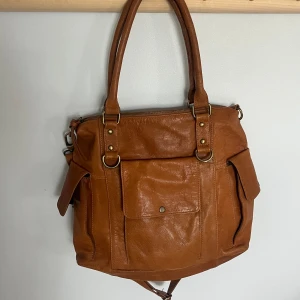 Brun axelväska i skinn från Wera - Snygg brun vintage axelväska från Wera i äkta skinn med flera fack, både med dragkedja och tryckknapp. Väskan har både handtag och avtagbar axelrem, samt detaljer i mässingsfärgad metall. Perfekt för dig som vill ha plats för allt du behöver under dagen. Inte använd så många gånger och har inga defekter och är i väldigt bra skick.