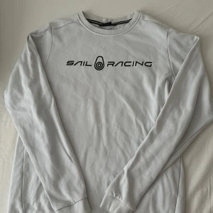 Vit Sail Racing sweatshirt - Vit sweatshirt från Sail Racing med svart logotyp tryckt över bröstet. Tröjan har rund hals, ribbade muddar och lång ärm. Materialet är mjuk bomull som känns skönt mot huden. Perfekt för dig som gillar stilren och sportig design.