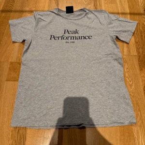 Grå Peak Performance t-shirt - Snygg grå t-shirt från Peak Performance med svart tryck på bröstet. Klassisk rund hals och korta ärmar, perfekt för chill dagar. Mjuk och skön bomull som känns nice mot huden. Enkel stil som funkar till jeans eller shorts.