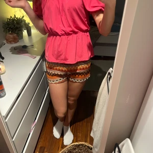 Rosa offshoulder blus med volang - Superfin rosa blus med offshoulder och puffiga korta ärmar. Blusen har resår i midjan och volang nertill, vilket ger en söt och trendig look. Perfekt för sommarens alla tillfällen och lätt att matcha med både jeans och kjol.