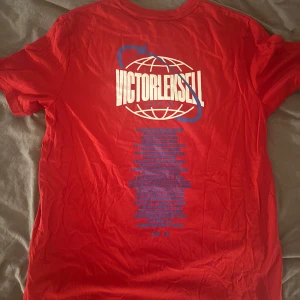 Röd Victor Leksell tour 2024 t-shirt M - Snygg röd t-shirt från Victor Leksell med tryck på ryggen från hans tour och patch med logga framtill. Klassisk rund hals och korta ärmar, tillverkad i mjuk bomull. Perfekt för dig som gillar merch och vill sticka ut med coolt tryck.