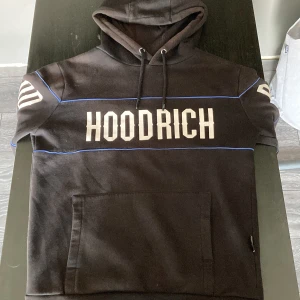 Svart HOODRICH hoodie med blå detaljer (St. XS/S) - Svart hoodie från HOODRICH med stor vit logga på bröstet och ärmarna. Hoodien har blå linjer som detaljer, huva med snörning och en stor magficka. Perfekt för en streetwear-look och chill dagar. Ops! Lite solbränd men inget som syns särskilt mycket. (St. XS/S)
