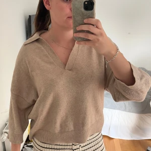 Beige stickad tröja med v-ringning och krage - Beige stickad tröja med avslappnad passform och v-ringning. Tröjan har en bred krage och trekvartsärmar, perfekt för lager-på-lager. Ribbstickade muddar vid ärmslut och nederkant ger en snygg detalj.