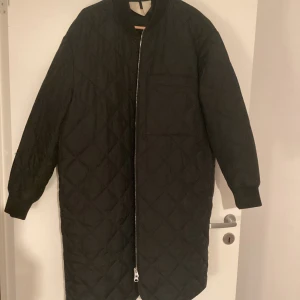 Svart quiltad lång jacka från ARKET Storlek M/L - Svart lång quiltad jacka från ARKET i storlek M/L. Jackan har ett stilrent rutmönster, lång ärm och dragkedja framtill. Perfekt för dig som gillar minimalistisk och clean stil. Materialet är polyester och jackan har en loose passform.