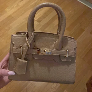 Beige handväska med gulddetaljer - Snygg beige handväska i skinn med guldfärgat spänne och dubbla handtag. Väskan har en stilren och klassisk design med struktur i materialet och avtagbar axelrem. Perfekt storlek för det viktigaste och passar till många outfits.