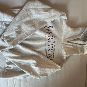 Grå hoodie Santa Catalina - Snygg ljusgrå hoodie från Gina tricot  med svart tryck framtill. Klassisk modell med huva och långa ärmar, perfekt för chill dagar. Mjuk bomullsblandning och ribbade muddar för extra komfort.