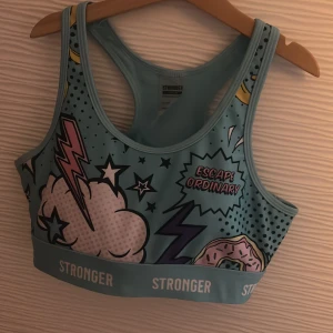 Blå sporttopp från Stronger med tryck - Cool blå sporttopp från Stronger med färgglatt serietidning-tryck, blixtar, stjärnor och text som 'Escape Ordinary'. Elastisk resår med logga, brottarrygg och håldetalj bak. Perfekt för dig som vill se cool ut på gymmet .