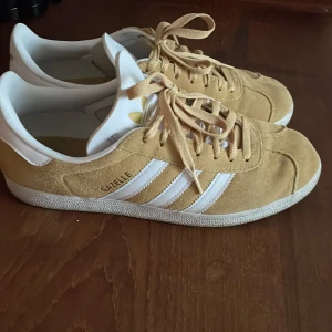 Adidas Gazelle beige mockasneakers - Snygga Adidas Gazelle sneakers i beige mocka med klassiska vita ränder och vit hälkappa. Skorna har snörning och platt sula, samt Adidas logga i guld på hälen och tungan. Perfekta för dig som gillar retrostil och vill sticka ut med en fräsch färg.