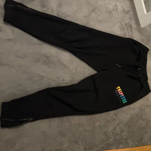 Svarta Trapstar mjukisbyxor med dragkedja - Svarta mjukisbyxor från Trapstar med färgglad logga på benet. Byxorna har snörning i midjan och dragkedjor vid bensluten. Perfekta för chill eller streetwear och tillverkade i mjukt bomullsmaterial. ♾️