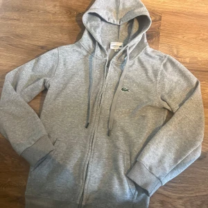 Grå hoodie från Lacoste med dragkedja - Snygg grå hoodie från lacoste. Säljer på grund av att den inte används. Tröjan är M men passar mer som xs/s. Hör av dig vid frågor 