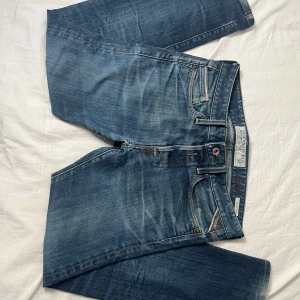 Replay Waitom blå jeans herr - Snygga blå jeans från Replay, modell Waitom. Klassisk femficksdesign med Replay-logga på myntfickan och röda detaljer vid midjan. Jeansen har en rak passform och är tillverkade i slitstarkt denimtyg. Perfekta för en avslappnad och cool stil. Säljes billigt pga litet litet håll vid skrevet