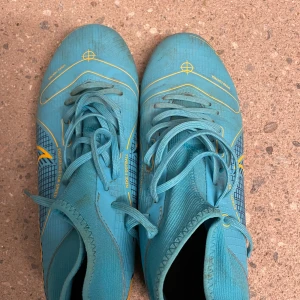 Blå Nike fotbollsskor med snörning - Säljer ett par blå Nike fotbollsskor med gula detaljer och snörning. Skorna har en smidig och sportig design med högre skaft och är tillverkade i syntetmaterial för bästa grepp och känsla på planen. Perfekta för dig som vill sticka ut på matchen!