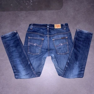 Nudie Jeans organic denim - Snygga blåa organic denim Nudie Jeans med klassisk femficksdesign och coola vågade sömmar på bakfickorna. Modellen är skinny med smal passform och har orangea kontrastsömmar samt Nudie-logga på läderpatchen bak. Perfekt för dig som gillar stilrena och moderna jeans 🤑‼️OBS har en lagning i skrevet ‼️