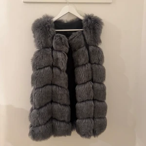 Grå fluffig pälsväst - Säljer en supermjuk och fluffig grå pälsväst med breda, markerade paneler. Västen är ärmlös och har en krage som ger extra volym. Perfekt för att lyfta vilken outfit som helst med en lyxig känsla och cool look.