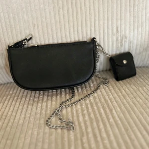 Svart axelväska med kedja från By Malene Birger - Snygg svart axelväska i skinn från By Malene Birger med silverfärgad kedjerem och matchande liten plånbok. Väskan har dragkedja upptill och stilren, halvmåneformad design. Perfekt för att bära det viktigaste och ge en cool touch till din outfit.