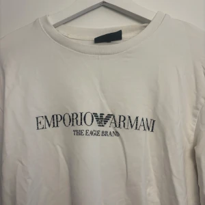 Vit t-shirt från Emporio Armani - Snygg vit tröja från Emporio Armani med klassisk rund hals och svart broderad logga framtill. T-shirten är i mjuk bomull och har en clean, stilren look som passar perfekt till jeans eller shorts.