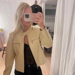 Beige skinnjacka med dragkedjor - Snygg beige skinnjacka från Gipsy med cool ståkrage och två zip-detaljer på bröstet. Jackan har en rak, lite croppad passform och långa ärmar. Perfekt t lite mer fest kläder.
