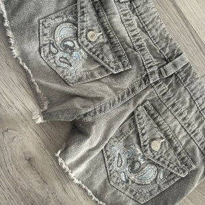 Grå jeansshorts med broderi Gina Tricot - Snygga grå jeansshorts från Gina Tricot med slitna kanter och coolt blått och vitt broderi på bakfickorna. Modellen är lågmidjad med klassiska fickor och silverfärgade knappar. Perfekt för dig som gillar detaljer och streetstyle. aldrig använda 