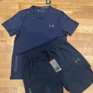 Under Armour set - Sportig blå t-shirt från Under Armour med korta ärmar och rund hals. T-shirten har ett stilrent tryck med loggan på bröstet och text längs sidan. Perfekt för dig som gillar att träna och vill ha en snygg och bekväm look.